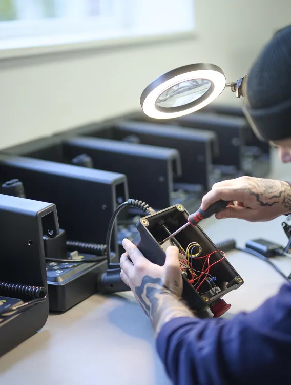 Technician working on a joystick control box with visible wiring, surrounded by similar units on the workbench.