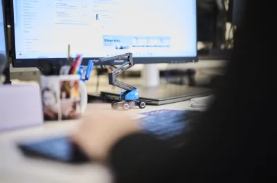 A close-up of an office desk with a computer screen, a miniature blue boom lift model, and a person typing on a keyboard.